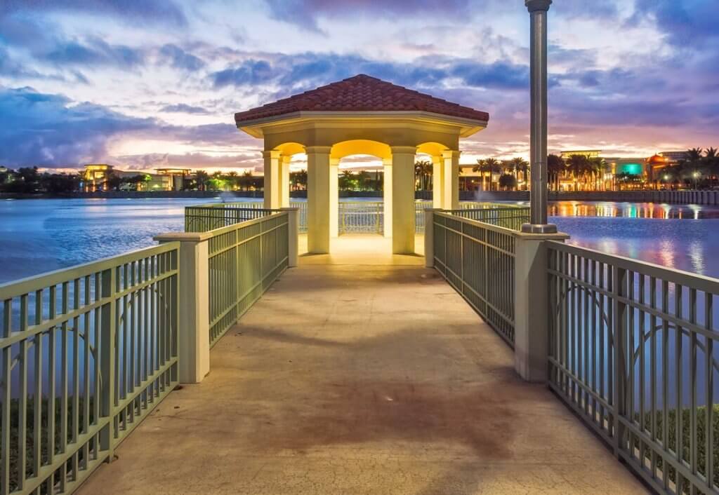 Eine heitere Abendszene mit einem Pavillon mit Ziegeldach am Ende eines Weges über einen ruhigen See, umgeben von einer Stadtlandschaft mit beleuchteten Gebäuden und Palmen
