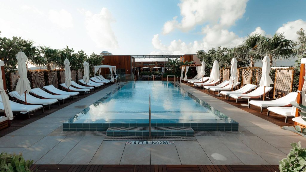 Uma luxuosa área de piscina na cobertura com cabanas brancas e espreguiçadeiras, cercada por uma vegetação exuberante e um céu azul claro