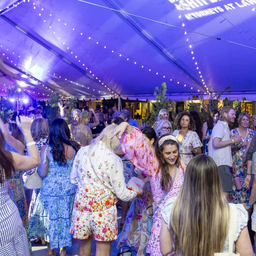 Une fête animée en plein air sous une grande tente blanche avec des guirlandes lumineuses colorées, où des invités vêtus de tenues d'été éclatantes dansent et se rencontrent.
