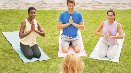 One man and two women in yoga postures on mats on lawn in front of female instructor