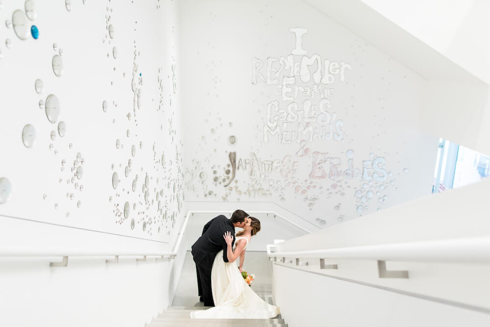 Bride and groom kissing on a staircase in front of a white wall of art