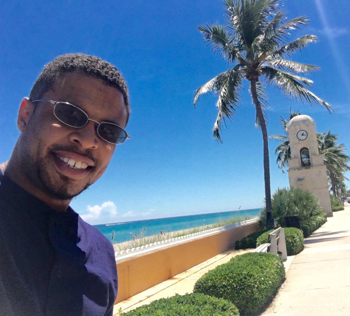 A young man in sunglasses takes a smiling selfie in front of the Worth Avenue clock tower