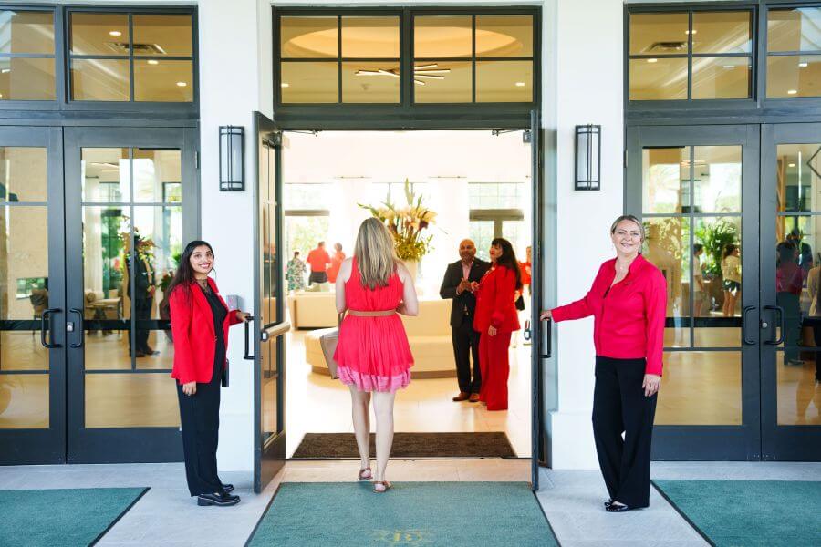 Three women in red outfits stand on either side of a grand entrance with glass doors, greeting guests arriving at an elegant event