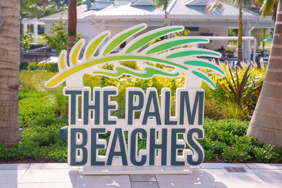 A vibrant sign with a palm leaf design reading 'The Palm Beaches' stands amidst lush greenery and palm trees
