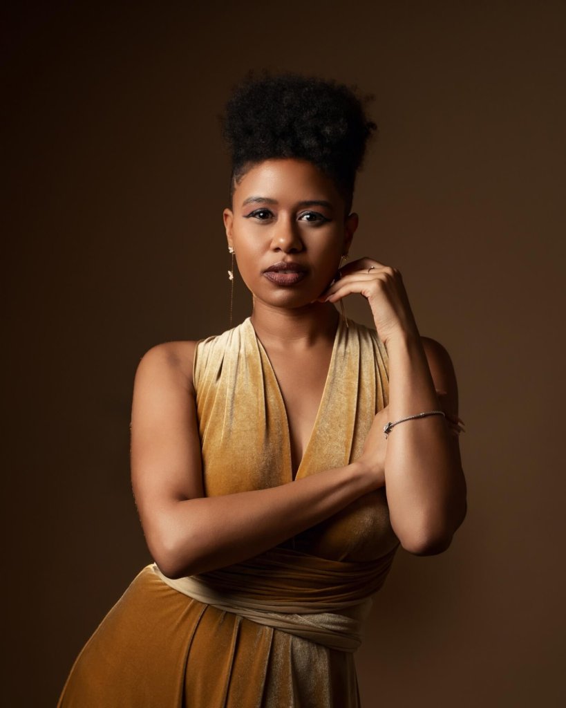 A woman with a confident expression, wearing a golden velvet dress and jewelry, stands with her arms crossed against a dark background