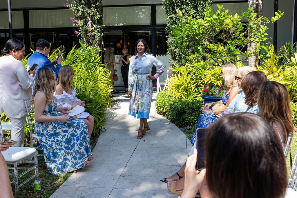 A woman in a colorful floral dress walks down a garden path lined with guests seated on either side, capturing the moment on their phones