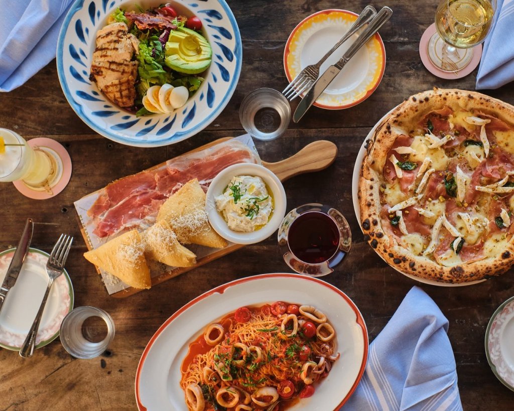 A rustic wooden table is set with an array of Italian dishes, including a grilled chicken salad, prosciutto and cheese platter, pizza with prosciutto and arugula, spaghetti with squid ink and calamari, and a variety of bread and dips, all accompanied by wine and water glasses
