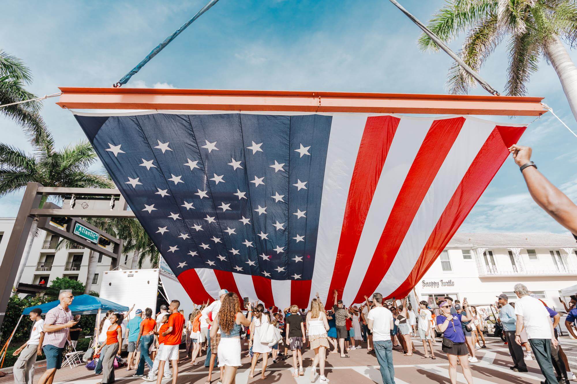 A large American flag is being held aloft by a crowd of people in a public square, with palm trees and buildings in the background