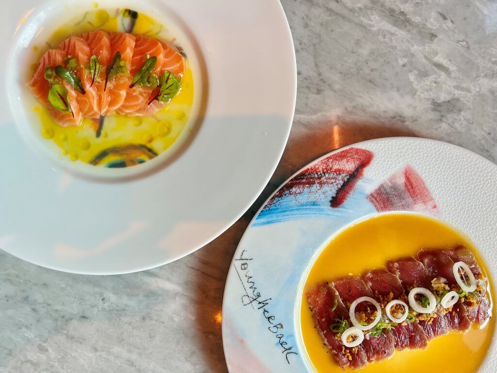 Two artistically plated dishes featuring raw fish: one with salmon slices on a yellow sauce, garnished with herbs; the other with tuna slices on a yellow sauce, topped with onion rings and herbs