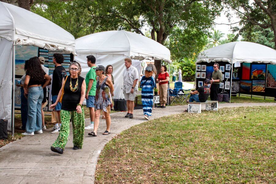 A lively outdoor art fair with white tents showcasing various artworks, surrounded by people browsing and enjoying the event under the shade of trees