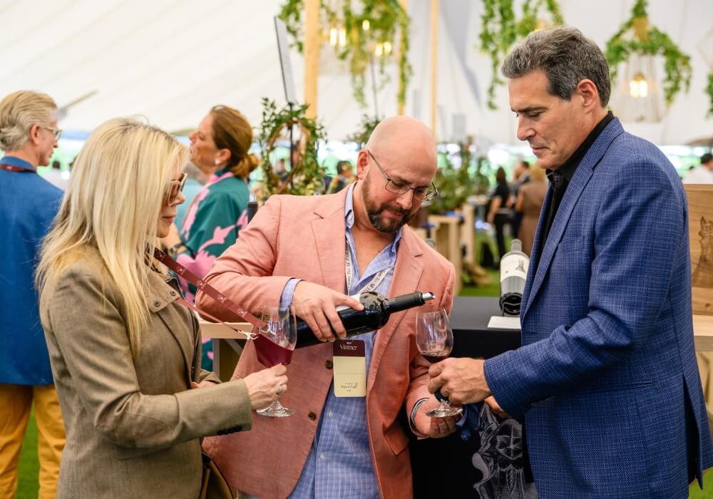 A man in a pink blazer pours red wine for people at an event