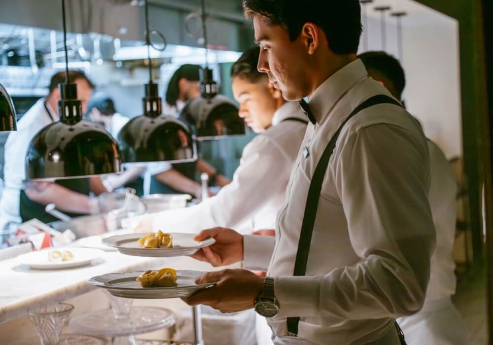 A waiter in a formal uniform holding plates of food in a restaurant kitchen