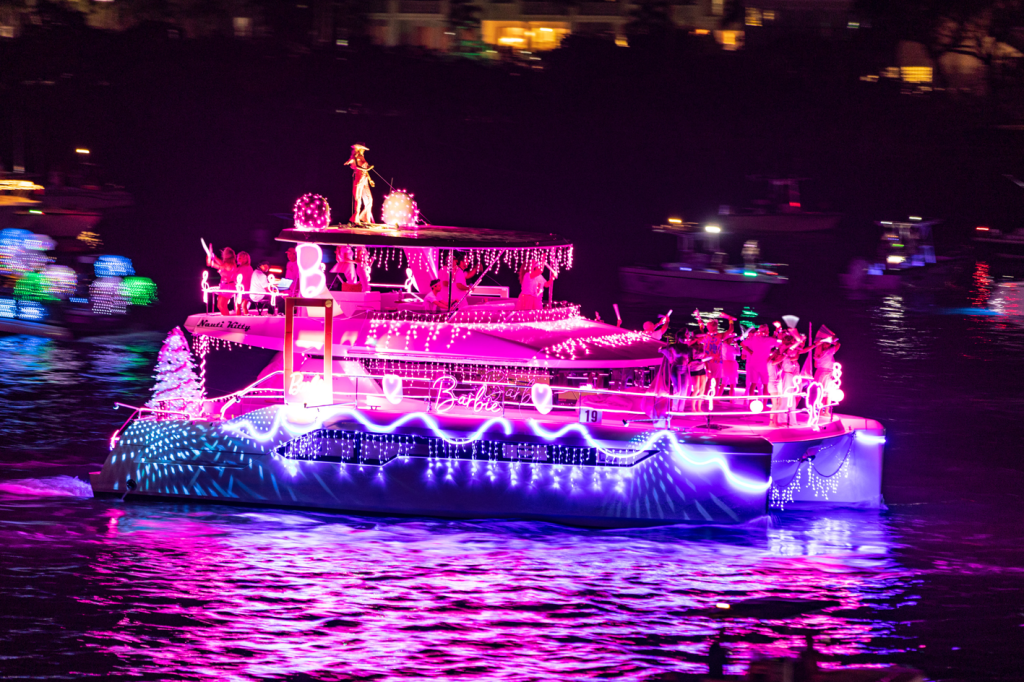 Un barco de color rosa decorado con luces y diseños inspirados en Barbie flota en el agua durante un desfile festivo nocturno. 