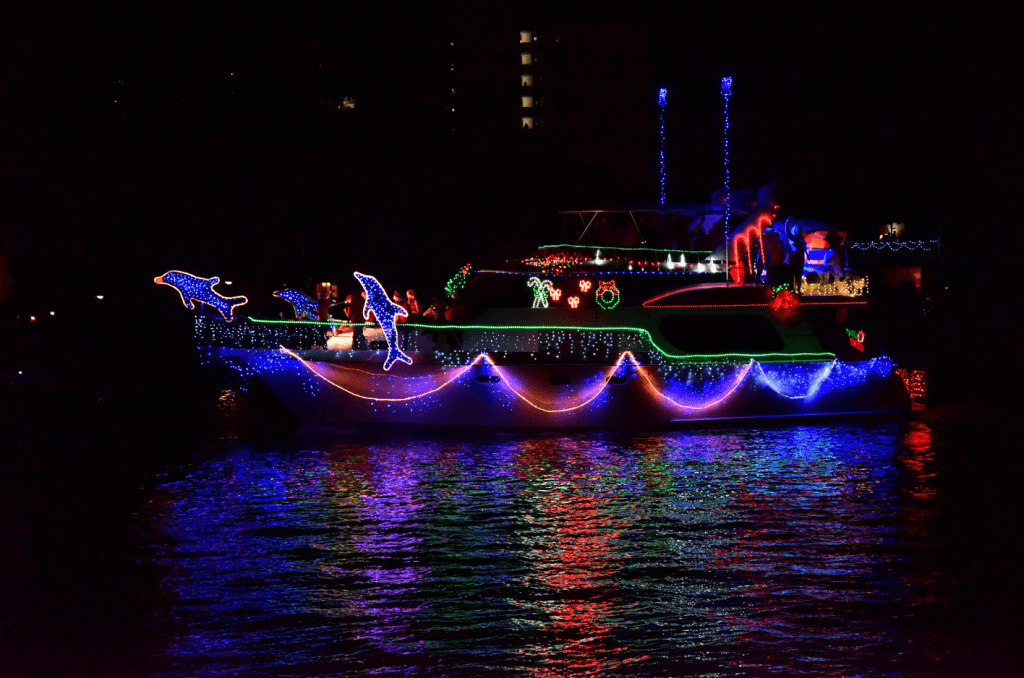 A festive boat parade featuring a brightly lit vessel adorned with colorful Christmas lights and decorations, including dolphins and holiday motifs