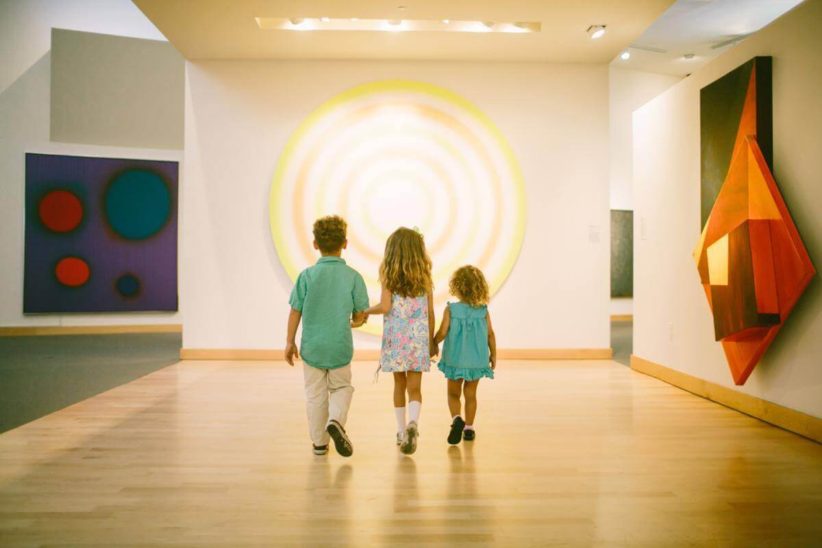 Drei Kinder gehen Hand in Hand durch eine moderne Kunstgalerie und bewundern bunte abstrakte Gemälde und ein großes rundes Kunstwerk an den Wänden