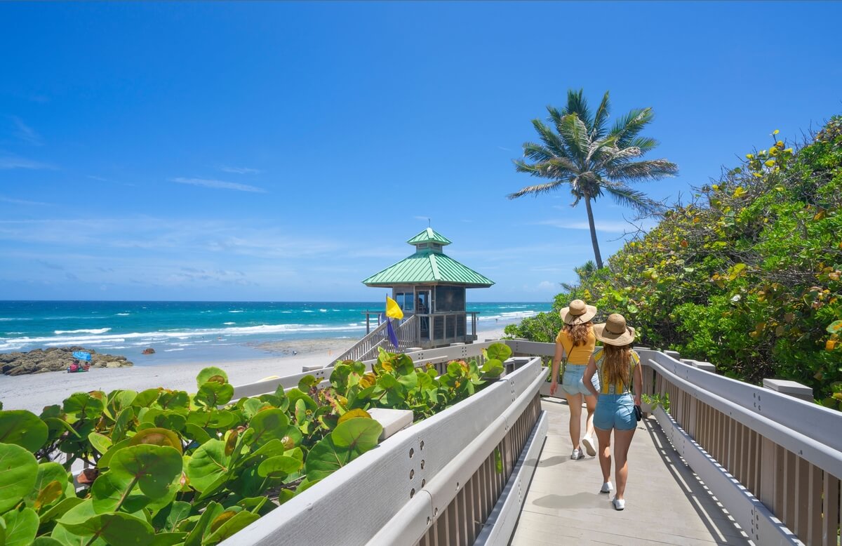 Dos mujeres con sombreros y ropa de verano caminan por una pasarela de madera bordeada de exuberantes plantas verdes hacia una pequeña glorieta verde cerca de la playa, con un cielo azul claro y el océano al fondo.