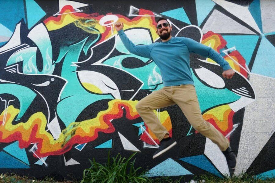Un hombre con camisa azul y gafas de sol se coloca delante de un llamativo mural de grafitis con formas abstractas y colores vivos