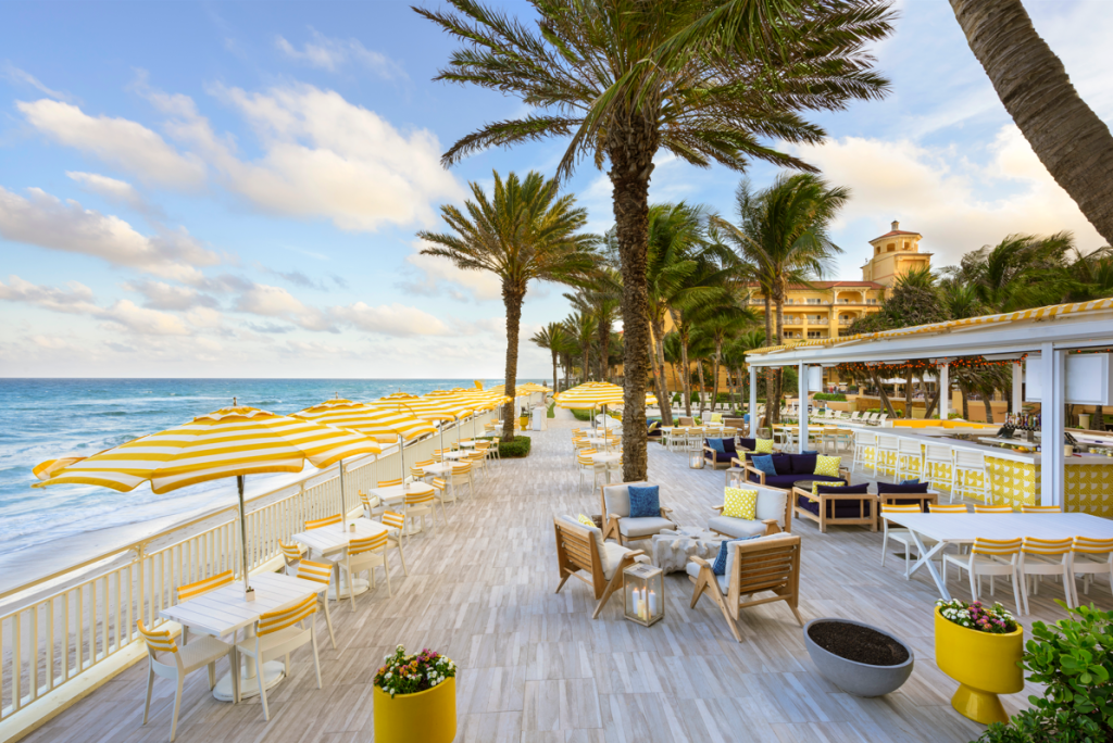 Outdoor dining area at Breeze Ocean Restaurant featuring yellow and white striped umbrellas, palm trees, and a  view of the ocean