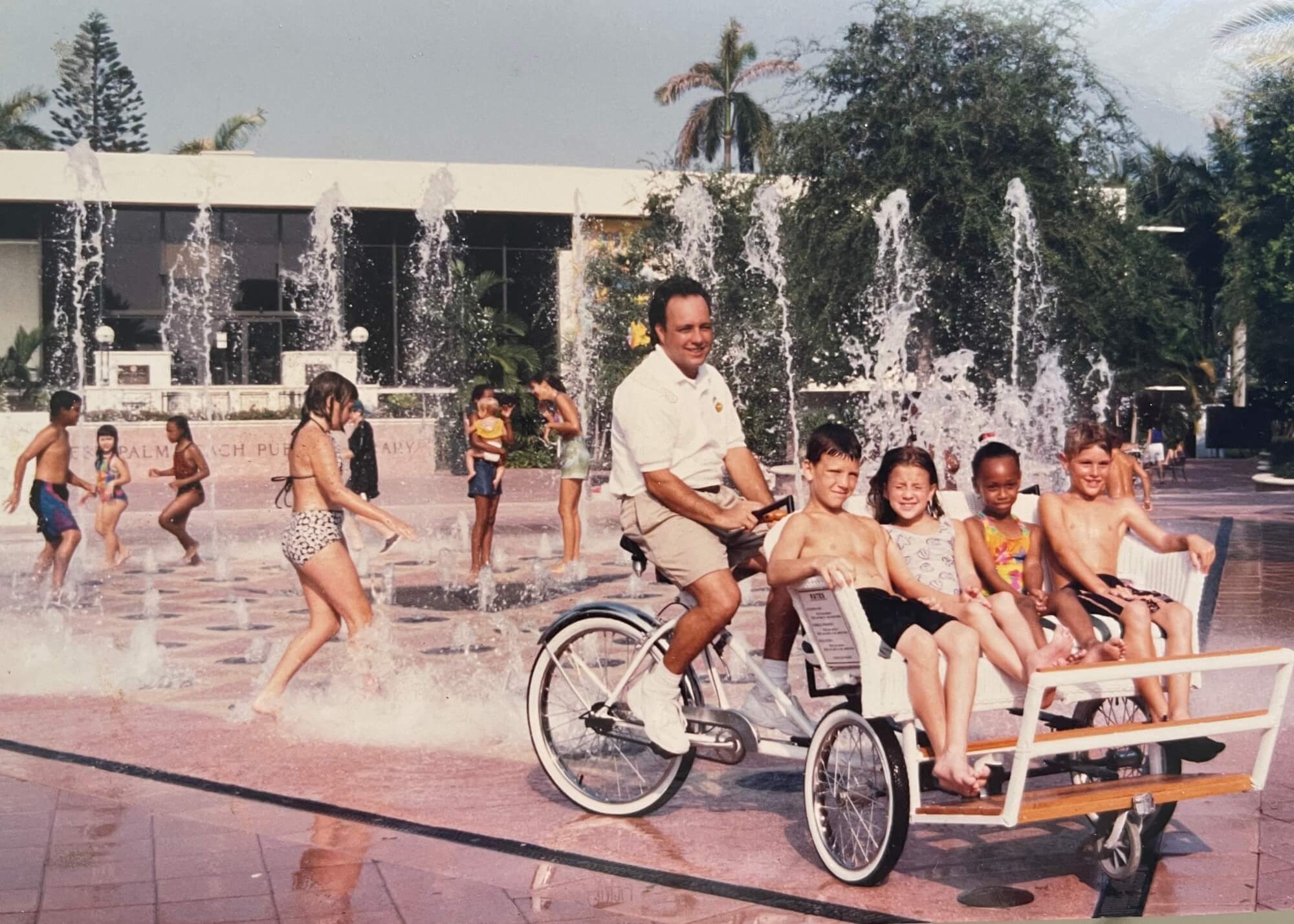 A man rides a multi-passenger bicycle past children playing in a public water fountain