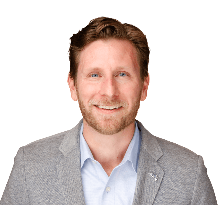 A man with short brown hair and a beard is smiling warmly at the camera, dressed in a light gray blazer over a light blue button-up shirt