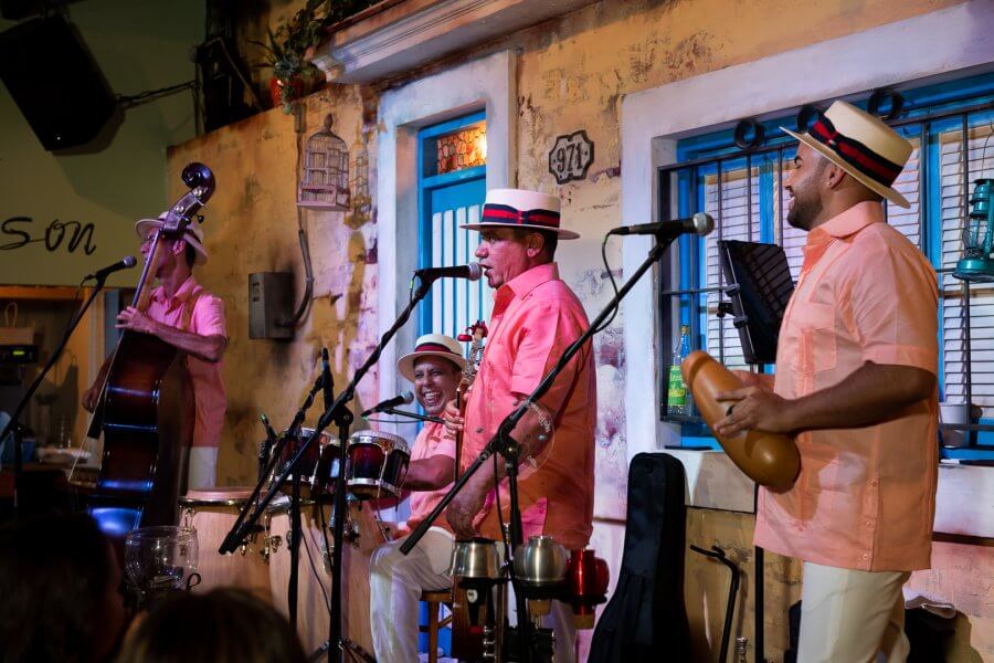 A lively band of four musicians dressed in matching pink shirts and white hats performs on a stage with a rustic backdrop, playing a variety of instruments including a double bass, drums, and percussion