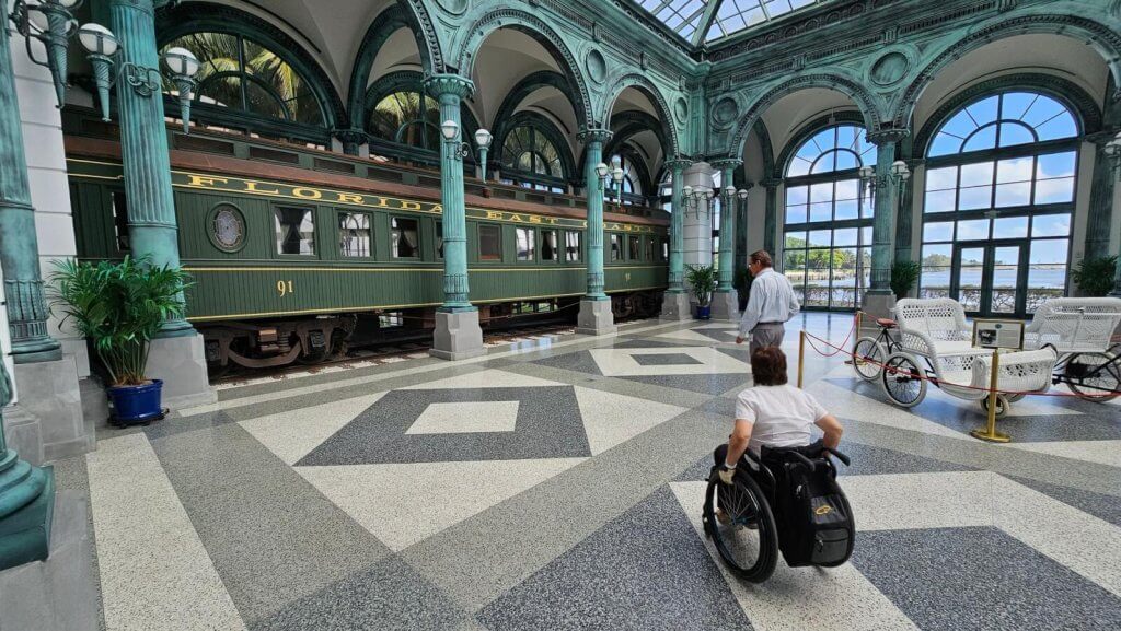 Uma pessoa em cadeira de rodas explora uma grande e histórica estação de trem com um vagão de trem antigo em exposição