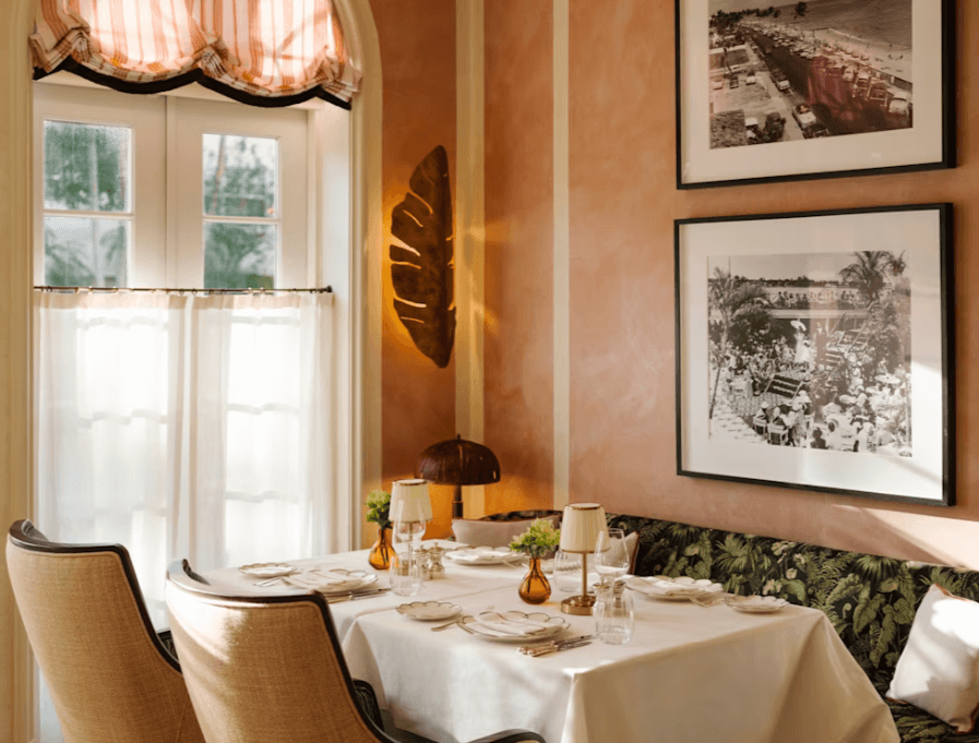 A tastefully decorated dining room with a tropical theme, featuring a table set for two with elegant tableware, framed black-and-white photographs on the walls, and a window with striped curtains allowing natural light to filter in