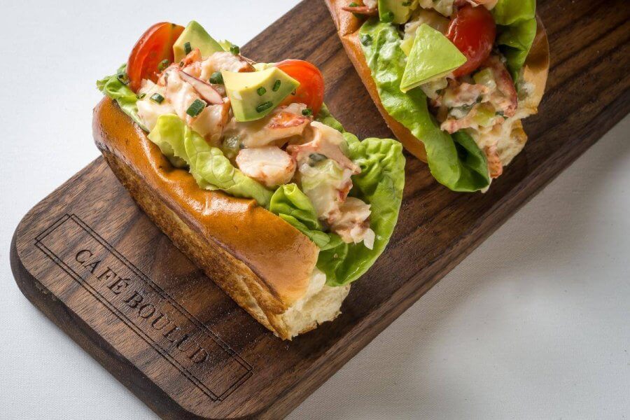 Two lobster rolls with lettuce, avocado, and tomatoes on a toasted bun, served on a wooden board