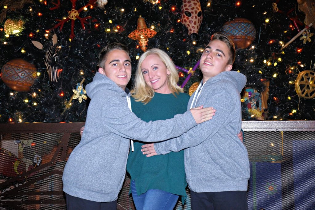 Three smiling individuals wearing gray hoodies embrace in front of a festive Christmas tree adorned with colorful ornaments and twinkling lights