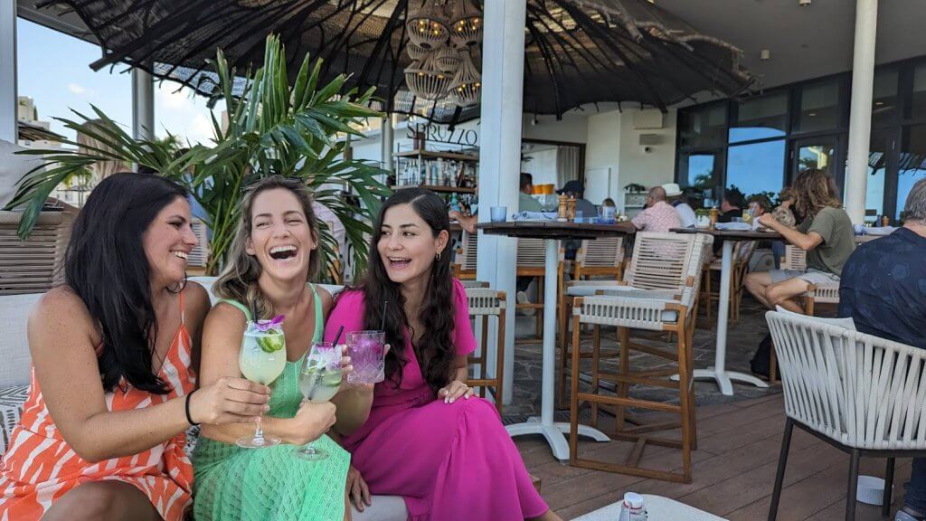 Tres mujeres con coloridos vestidos de verano ríen y disfrutan de unos cócteles juntas en un restaurante al aire libre con ambiente tropical