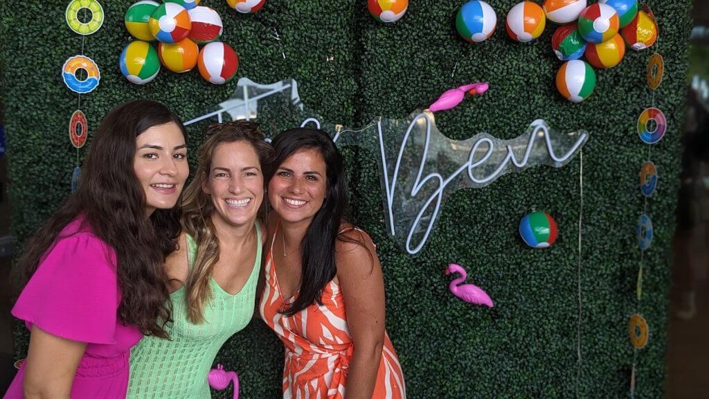 Tres mujeres con coloridos vestidos de verano posan juntas frente a un fondo festivo con un letrero de neón, pelotas de playa y un flamenco