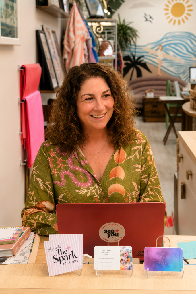 A woman with curly hair smiles warmly behind a desk with colorful business cards and a laptop