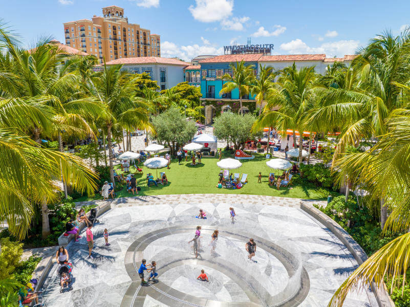 A vibrant outdoor plaza bustling with people enjoying a sunny day, surrounded by lush palm trees and colorful buildings