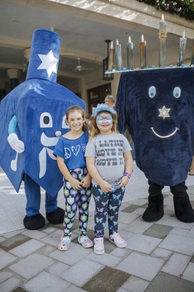 Two young girls stand between two large, friendly-looking characters dressed as a dreidel and a menorah, all smiling and posing for a photo at a Hanukkah celebration