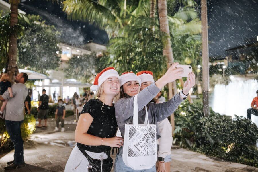 Three young women in Santa hats take a selfie amidst falling snow in a tropical setting, surrounded by palm trees and a festive crowd