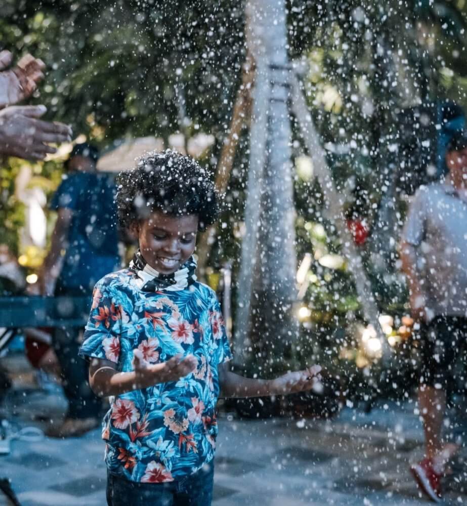 A joyful child in a colorful floral shirt delights in the falling snow, hands outstretched, surrounded by a festive crowd and lush greenery
