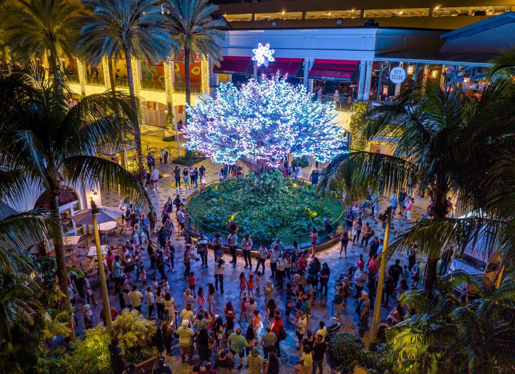 A vibrant nighttime scene in a tropical shopping center courtyard, featuring a large illuminated tree with blue lights, surrounded by palm trees and a crowd of people enjoying the festive atmosphere