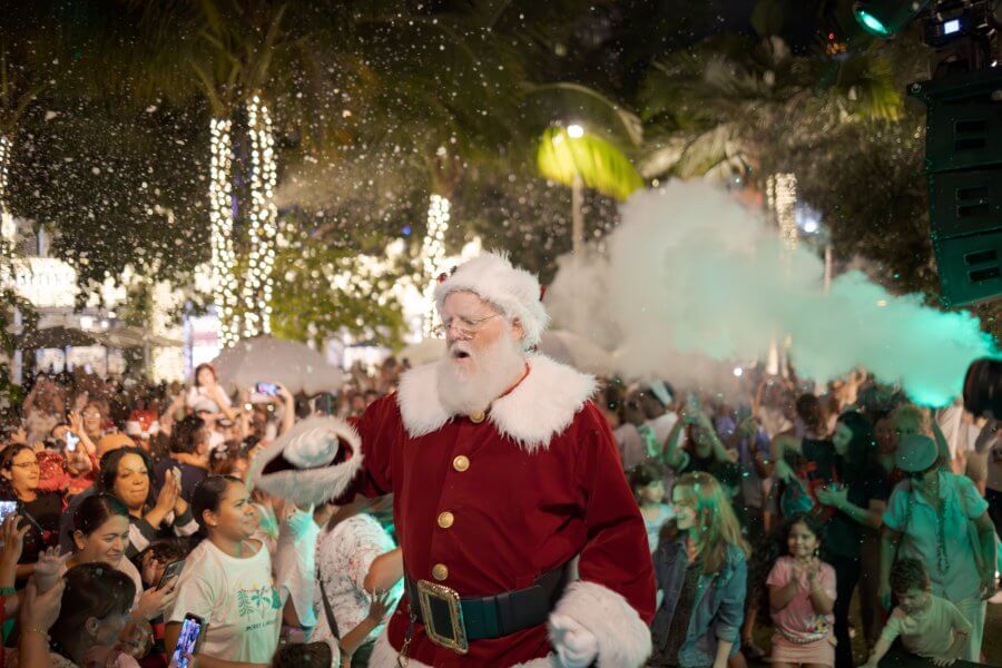 Santa Claus joyfully greets a cheering crowd amidst a festive parade with confetti and lights