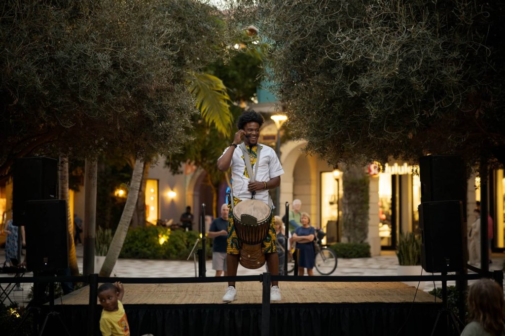 A musician performs on stage with a traditional drum in a lively outdoor setting surrounded by trees and onlookers