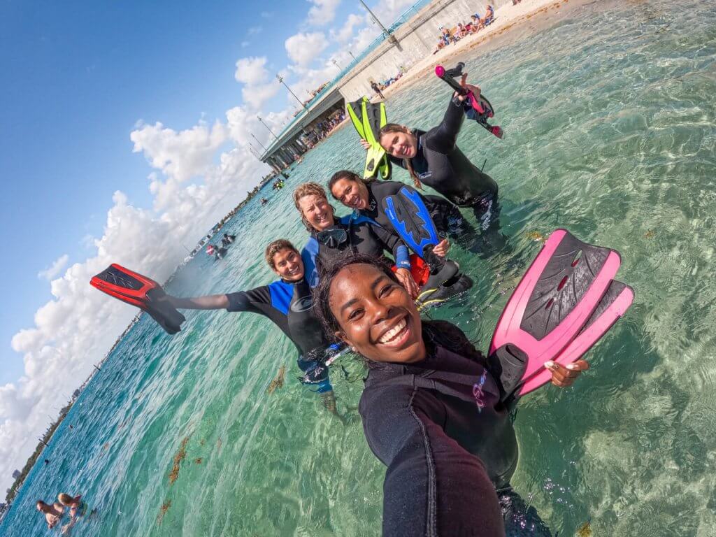 A group of five people in wetsuits and snorkel gear pose for a selfie in clear, shallow ocean water with a beach and bridge in the background