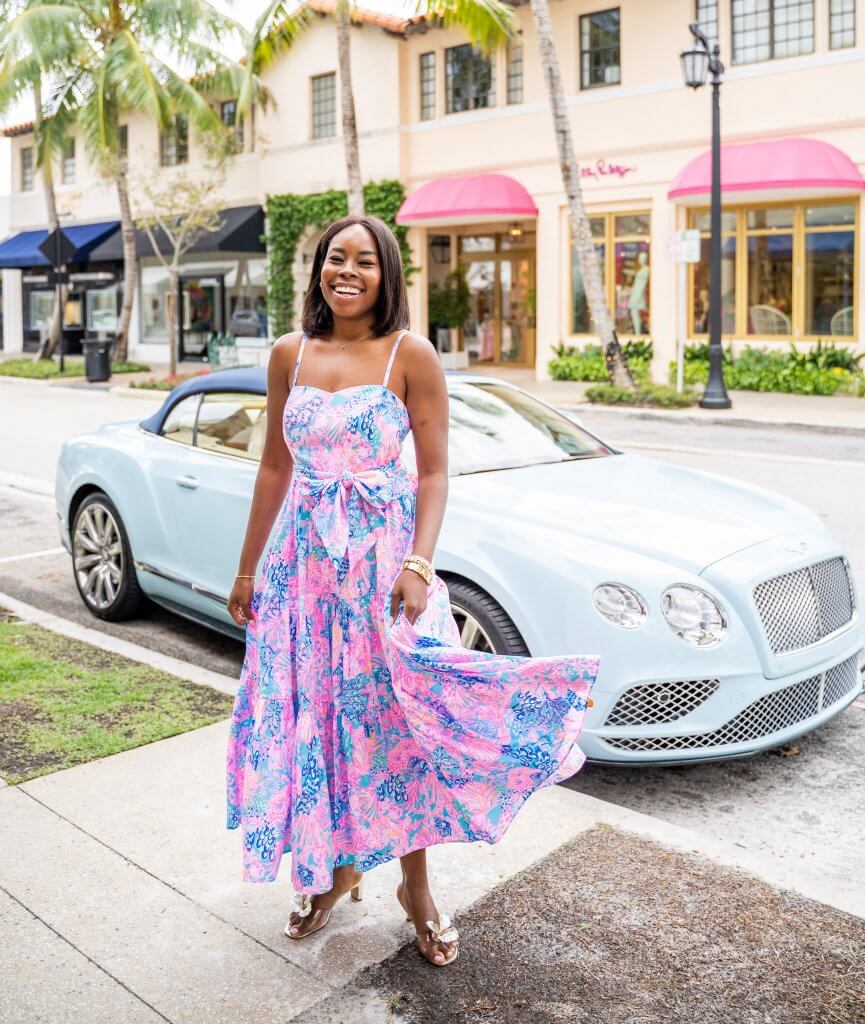 A woman in a vibrant floral dress stands on a sidewalk next to a light blue convertible car, with palm trees and pastel-colored buildings in the background