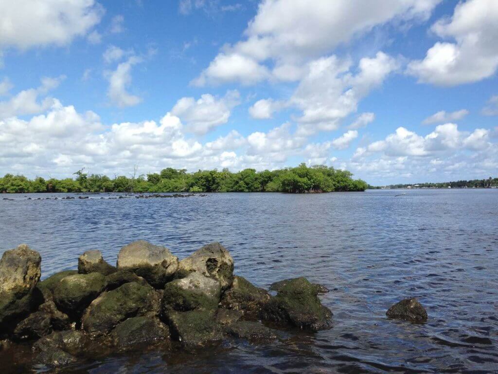 Une scène côtière sereine avec un rivage rocheux, des eaux calmes et des îles verdoyantes sous un ciel bleu vif parsemé de nuages blancs et floconneux.
