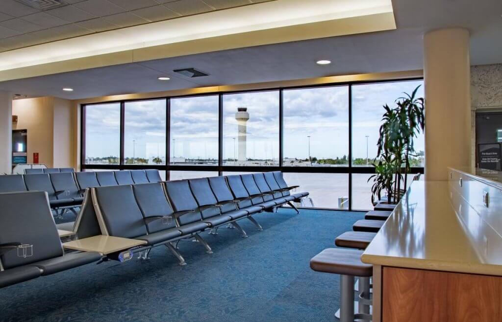 Una amplia terminal de aeropuerto con filas de asientos de cuero negro, un largo mostrador con taburetes y grandes ventanales que ofrecen vistas a la pista y a la torre de control