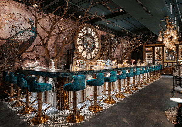 An ornate bar with teal velvet stools, a large clock, and peacock wall art