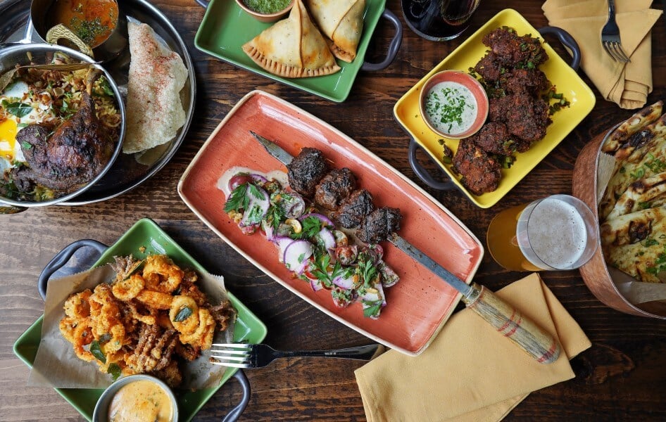 A vibrant spread of Indian and Middle Eastern dishes, including kebabs, fried snacks, curries, and bread, accompanied by colorful sauces and a glass of beer