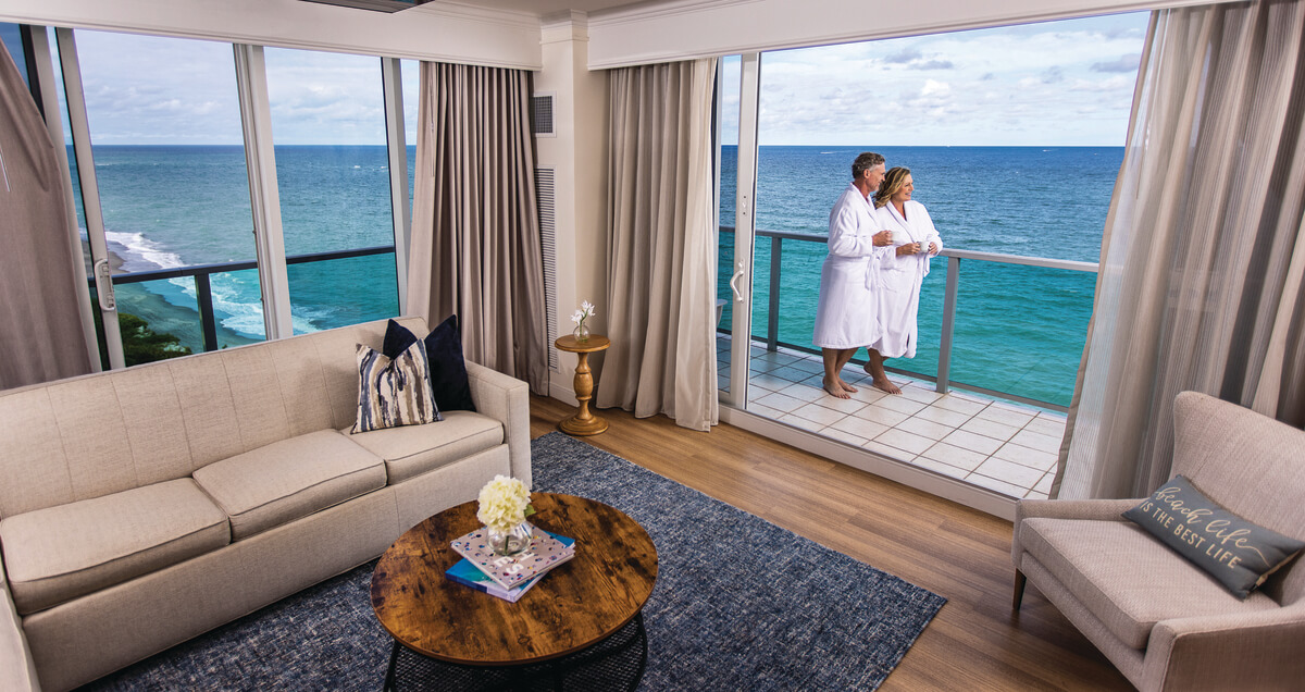 A couple in white robes stands on a balcony overlooking the ocean