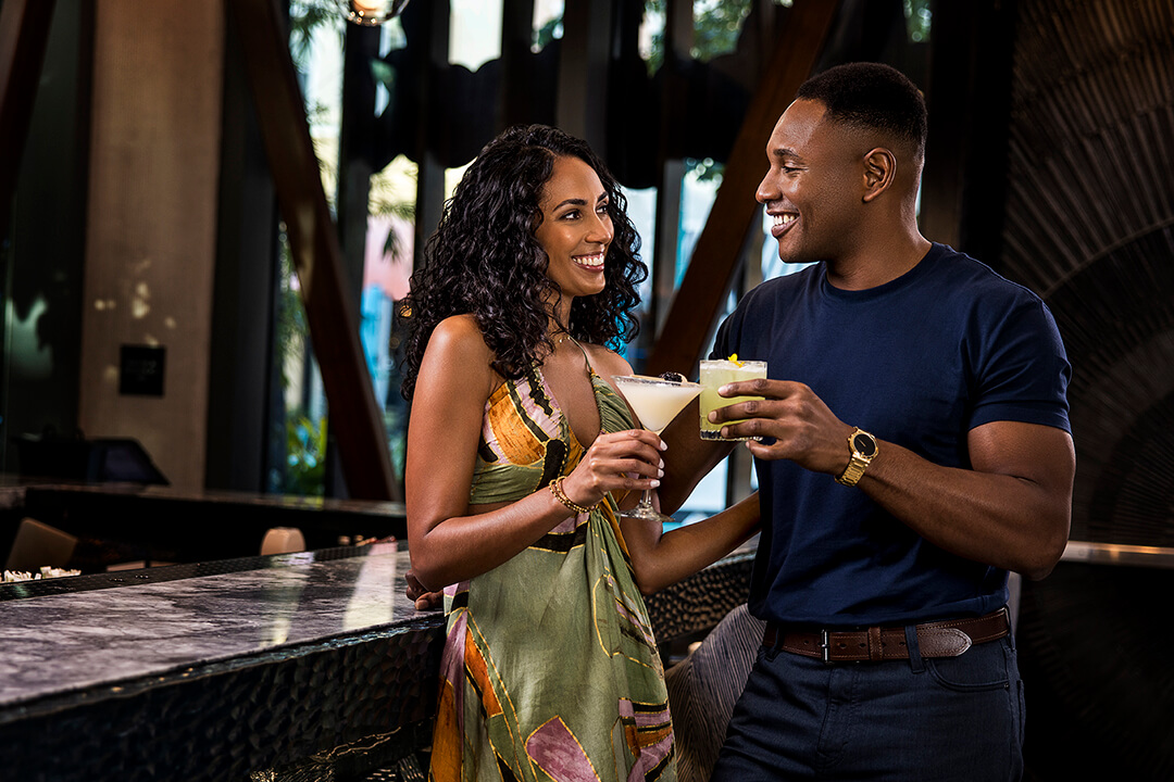 Un couple déguste des cocktails dans un bar élégant. La femme porte une robe vibrante et l'homme une chemise marine, tous deux sourient et trinquent.