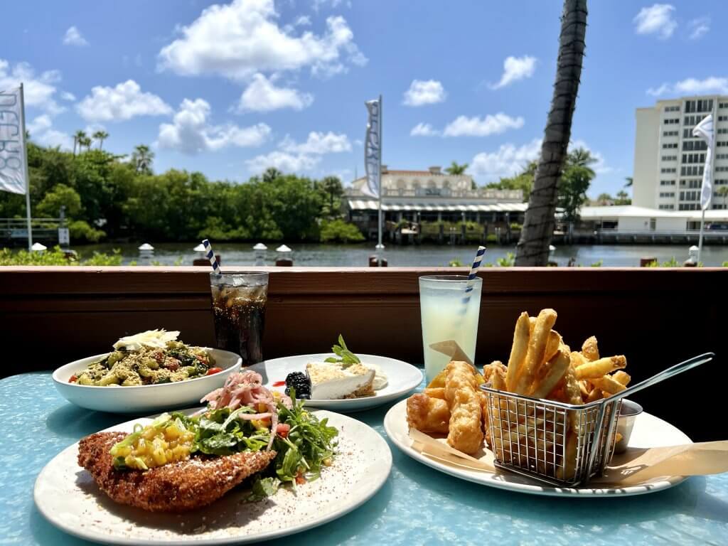 Se sirve una gran variedad de platos, como pollo frito, ensaladas y pescado con papas fritas, en una mesa con vistas a un pintoresco paseo marítimo con palmeras y edificios al fondo