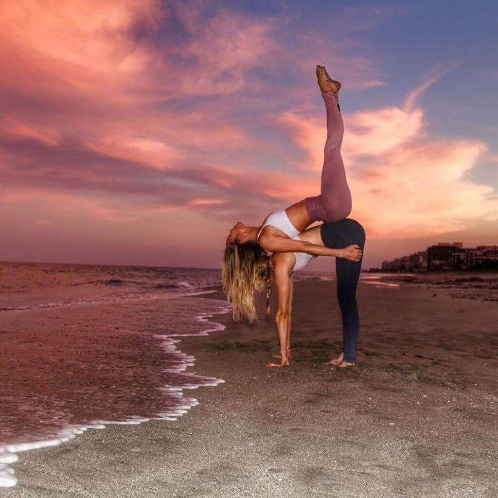 Dos mujeres realizando una postura de yoga sincronizada en una playa al atardecer, con una de ellas haciendo equilibrio sobre la espalda de la otra