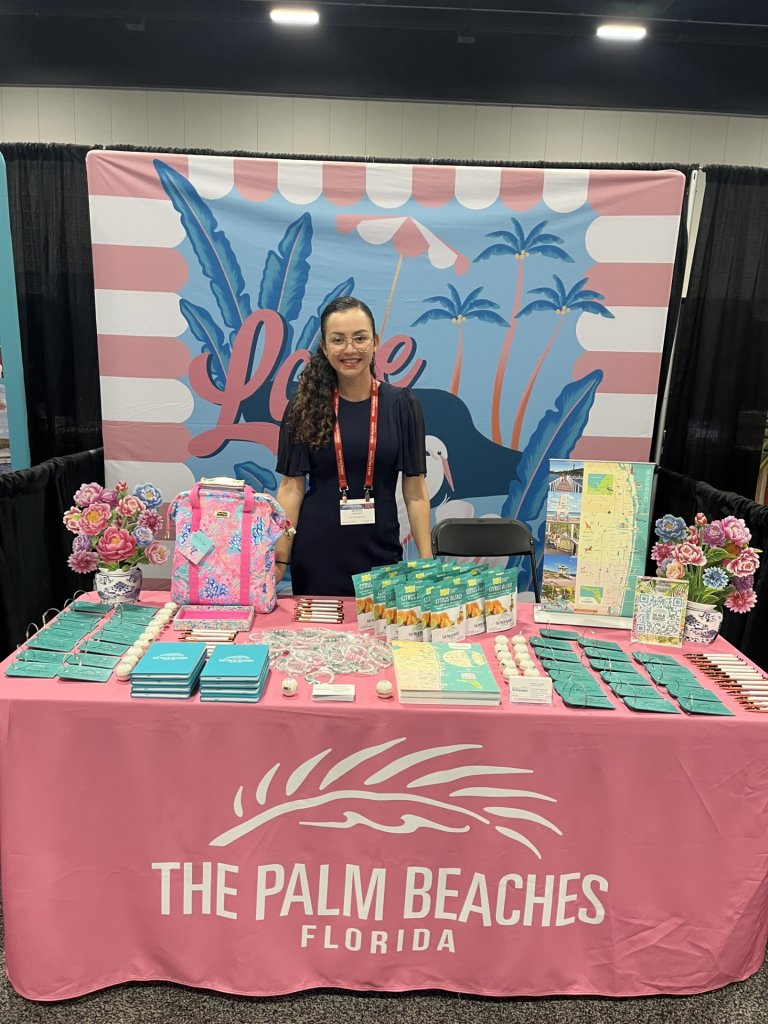 A woman stands behind a branded table for The Palm Beaches at a trade show